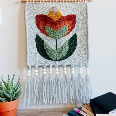 Zdjęcie produktu. Makatka z kwiatem w stylu folkowym. Na szarym tle czerwono-pomarańczowy kwiat. Ozdobiona długimi frędzlami, zawieszona na kiju.