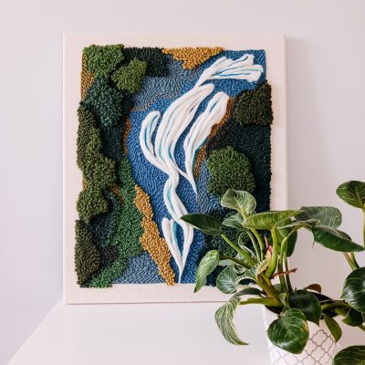 Teksturowy haft wykonany techniką punch needle, przedstawiający abstrakcyjny krajobraz z płynącą rzeką, otoczoną zielonymi drzewami i brązowymi elementami symbolizującymi ziemię. Obraz na jasnej kanwie stoi na białej powierzchni przy szarej ścianie. Obok niego znajduje się roślina doniczkowa o dużych, zielonych liściach w białej doniczce z geometrycznym wzorem.