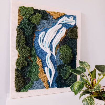 Teksturowy haft wykonany techniką punch needle, przedstawiający abstrakcyjny krajobraz z płynącą rzeką, otoczoną zielonymi drzewami i brązowymi elementami symbolizującymi ziemię. Obraz na jasnej kanwie stoi na białej powierzchni przy szarej ścianie. Obok niego znajduje się roślina doniczkowa o dużych, zielonych liściach w białej doniczce z geometrycznym wzorem.