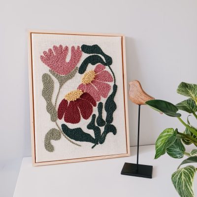 Haft wykonany techniką punch needle, przedstawiający różowe i bordowe kwiaty z żółtymi środkami oraz zielonymi liśćmi na lnianym tle, oprawiony w jasnobrązową drewnianą ramkę. Obraz stoi na białej powierzchni przy jasnoszarej ścianie. Obok niego znajduje się drewniana figurka ptaka na czarnym metalowym stojaku oraz kwiat doniczkowy.
