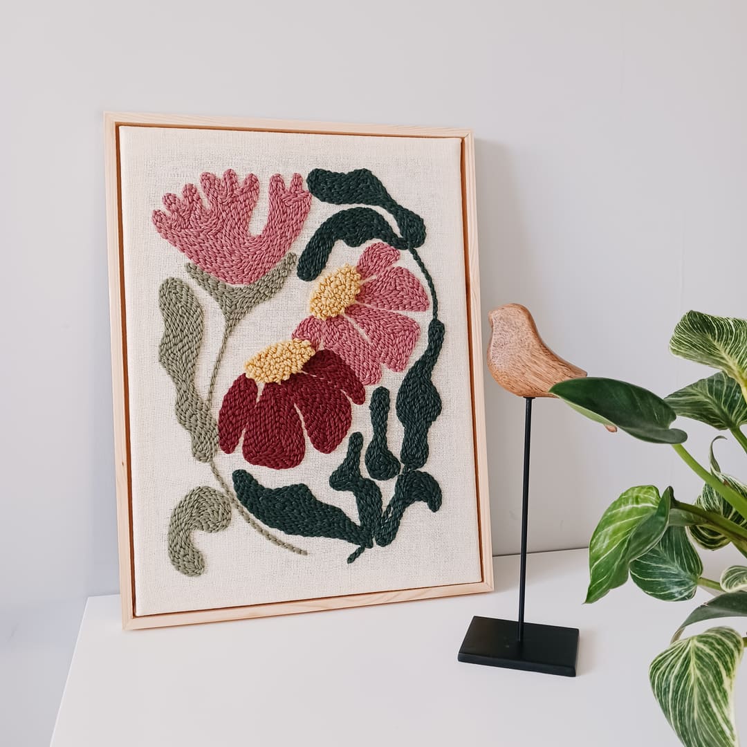 Haft wykonany techniką punch needle, przedstawiający różowe i bordowe kwiaty z żółtymi środkami oraz zielonymi liśćmi na lnianym tle, oprawiony w jasnobrązową drewnianą ramkę. Obraz stoi na białej powierzchni przy jasnoszarej ścianie. Obok niego znajduje się drewniana figurka ptaka na czarnym metalowym stojaku oraz kwiat doniczkowy.