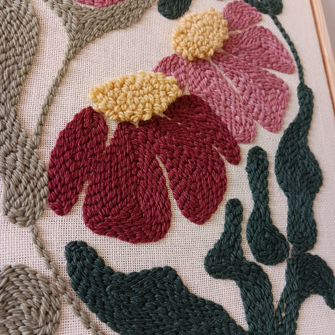 Fragment haftu wykonanego techniką punch needle, przedstawiający różowe i bordowe kwiaty z żółtymi środkami oraz zielonymi liśćmi na lnianym tle.