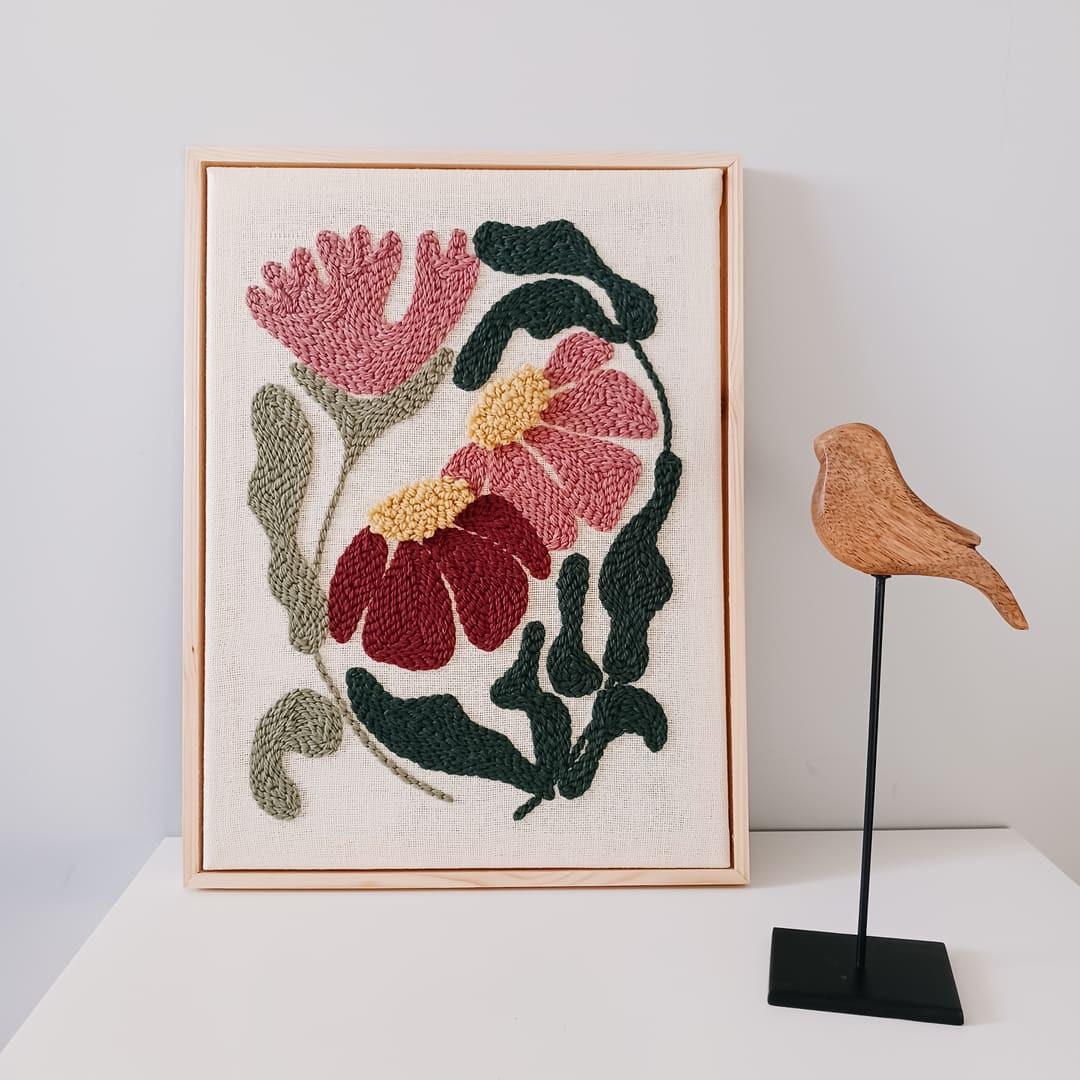 Haft wykonany techniką punch needle, przedstawiający różowe i bordowe kwiaty z żółtymi środkami oraz zielonymi liśćmi na lnianym tle, oprawiony w jasnobrązową drewnianą ramkę. Obraz stoi na białej powierzchni przy jasnoszarej ścianie. Obok niego znajduje się drewniana figurka ptaka na czarnym metalowym stojaku.