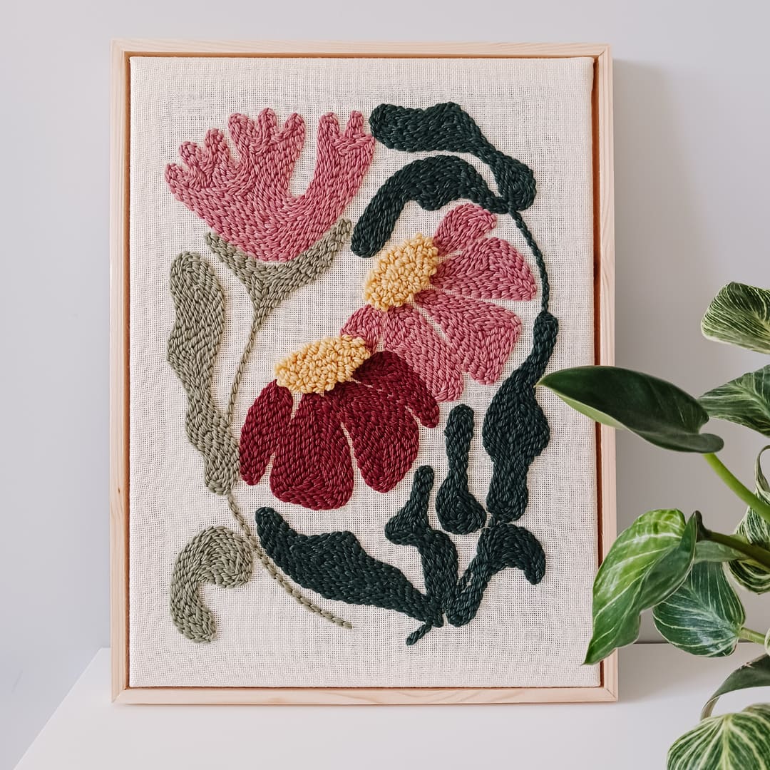 Haft wykonany techniką punch needle, przedstawiający różowe i bordowe kwiaty z żółtymi środkami oraz zielonymi liśćmi na lnianym tle, oprawiony w jasnobrązową drewnianą ramkę. Obraz stoi na białej powierzchni przy jasnoszarej ścianie. Obok niego znajduje się kwiat doniczkowy.