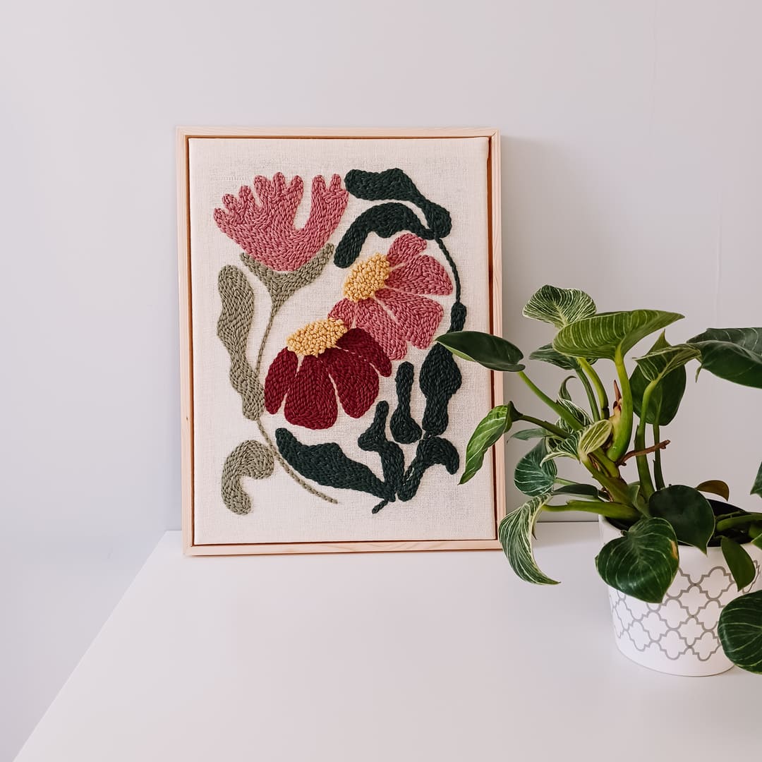 Haft wykonany techniką punch needle, przedstawiający różowe i bordowe kwiaty z żółtymi środkami oraz zielonymi liśćmi na lnianym tle, oprawiony w jasnobrązową drewnianą ramkę. Obraz stoi na białej powierzchni przy jasnoszarej ścianie. Obok niego znajduje się kwiat doniczkowy.