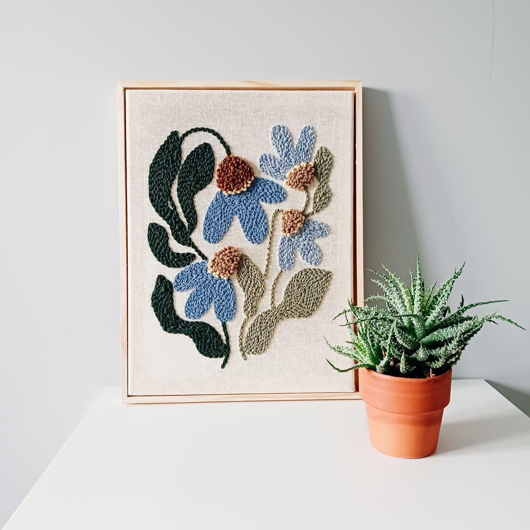 Haft wykonany techniką punch needle przedstawiający niebieskie kwiaty z brązowymi środkami oraz zielonymi liśćmi na lnianym tle, oprawiony w jasnobrązową drewnianą ramkę. Obraz stoi na białej powierzchni przy szarej ścianie. Obok niego znajduje się doniczka z zieloną rośliną, przypominającą sukulent, w glinianej doniczce.