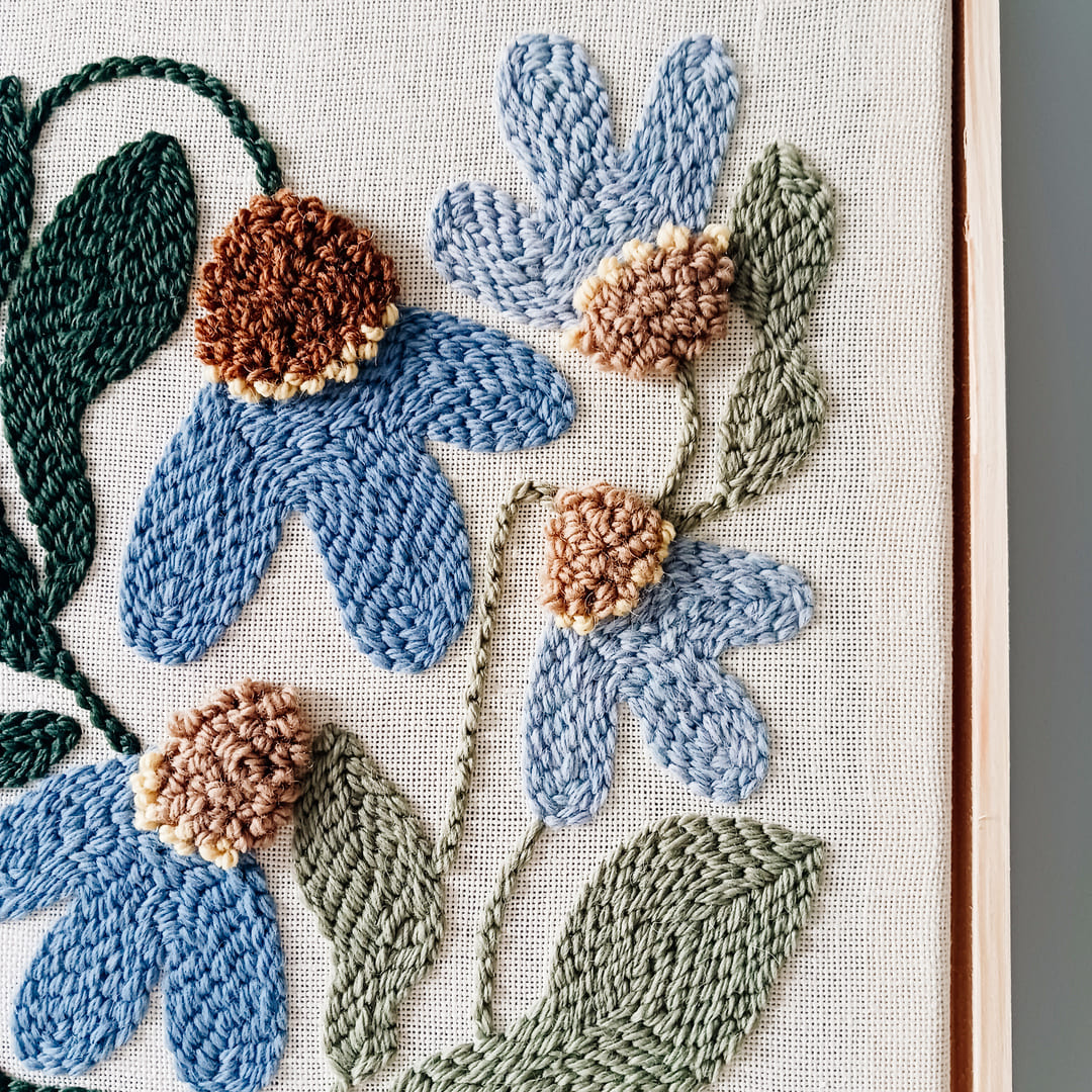 Fragment haftu wykonanego techniką punch needle przedstawiający niebieskie kwiaty z brązowymi środkami na lnianym tle. Z prawej strony widoczny kawałek ramki z surowego jasnego drewna.