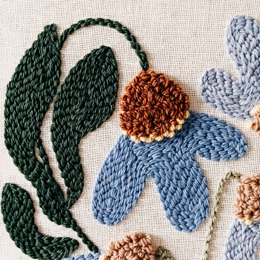 Fragment haftu wykonanego techniką punch needle przedstawiający niebieski kwiat z brązowym środkiem na lnianym tle.