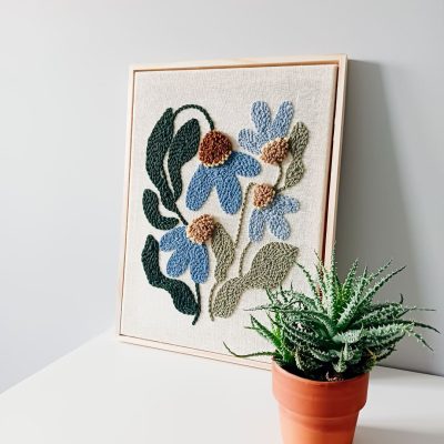 Haft wykonany techniką punch needle przedstawiający niebieskie kwiaty z brązowymi środkami oraz zielonymi liśćmi na lnianym tle, oprawiony w jasnobrązową drewnianą ramkę. Obraz stoi na białej powierzchni przy szarej ścianie. Obok niego znajduje się doniczka z zieloną rośliną, przypominającą sukulent, w glinianej doniczce.