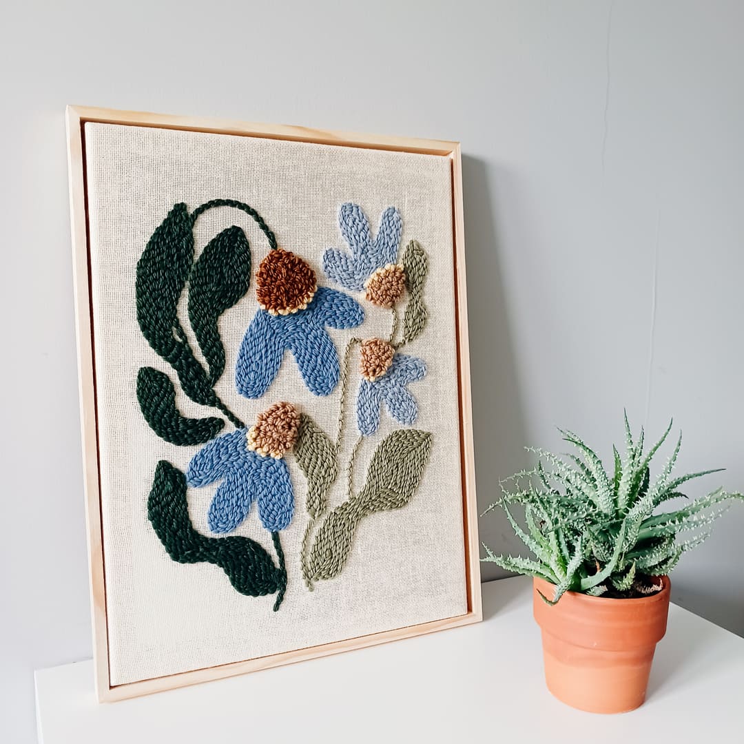 Haft wykonany techniką punch needle przedstawiający niebieskie kwiaty z brązowymi środkami oraz zielonymi liśćmi na lnianym tle, oprawiony w jasnobrązową drewnianą ramkę. Obraz stoi na białej powierzchni przy szarej ścianie. Obok niego znajduje się doniczka z zieloną rośliną, przypominającą sukulent, w glinianej doniczce.