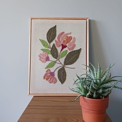 Haft wykonany techniką punch needle, przedstawiający różowe kwiaty z zielonymi liśćmi na lnianym tle, oprawiony w jasnobrązową drewnianą ramkę. Obraz stoi na drewnianej powierzchni przy jasnoszarej ścianie. Obok niego znajduje się mały sukulent w glinianej doniczce.