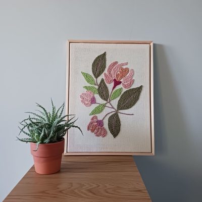 Haft wykonany techniką punch needle, przedstawiający różowe kwiaty z zielonymi liśćmi na lnianym tle, oprawiony w jasnobrązową drewnianą ramkę. Obraz stoi na drewnianej powierzchni przy jasnoszarej ścianie. Obok niego znajduje się mały sukulent w glinianej doniczce.