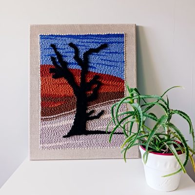 Dekoracyjny obraz wykonany techniką punch needle, przedstawiający czarne drzewo na tle pustynnego krajobrazu z czerwonymi wydmami i niebieskim niebem. Obraz oparty o ścianę, ustawiony na białej powierzchni obok doniczki z zieloną rośliną.