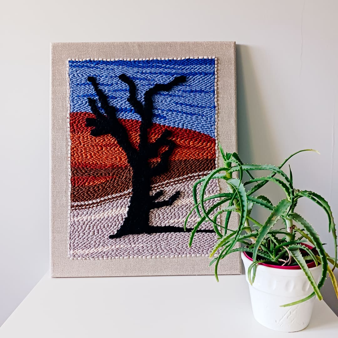 Dekoracyjny obraz wykonany techniką punch needle, przedstawiający czarne drzewo na tle pustynnego krajobrazu z czerwonymi wydmami i niebieskim niebem. Obraz oparty o ścianę, ustawiony na białej powierzchni obok doniczki z zieloną rośliną.