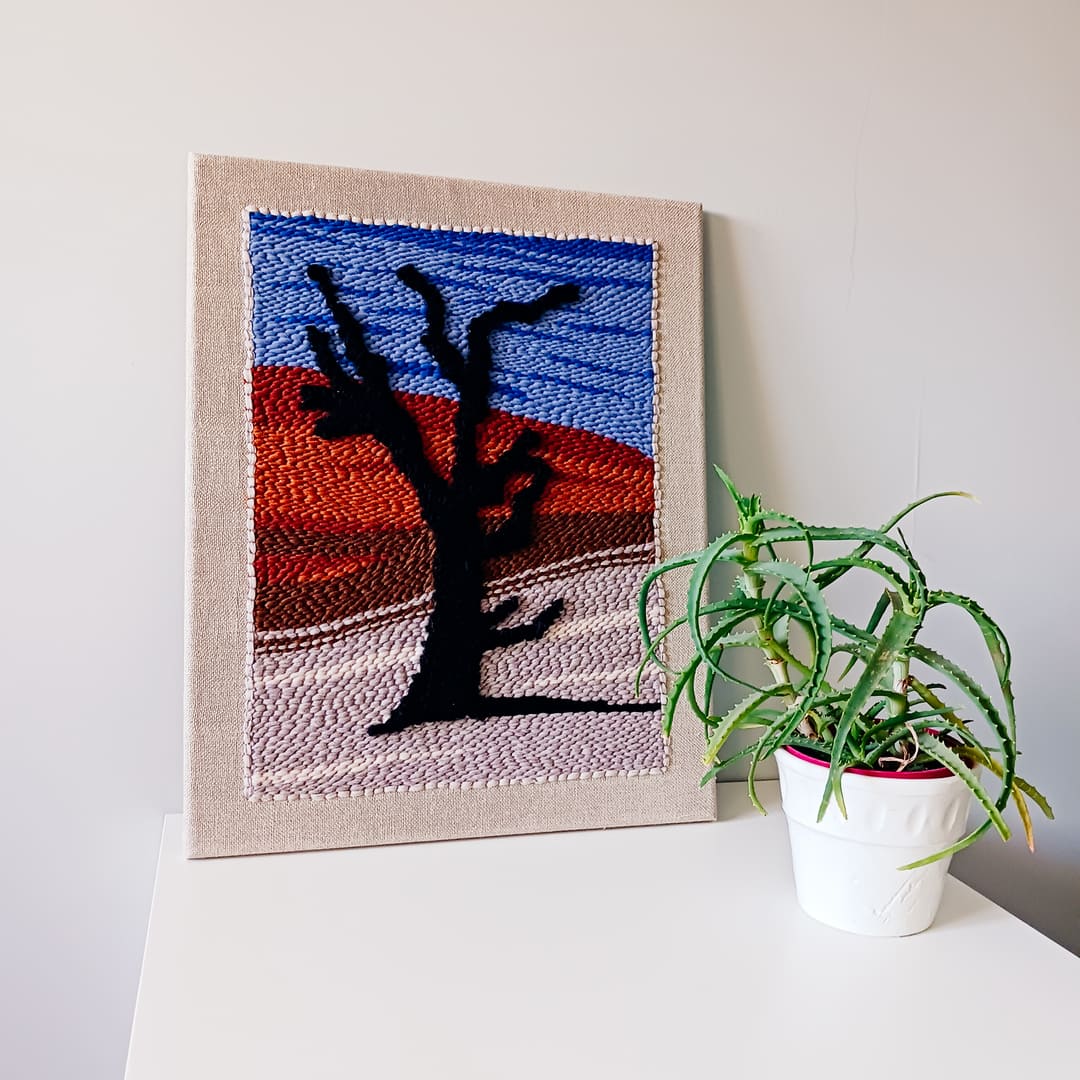 Dekoracyjny obraz wykonany techniką punch needle, przedstawiający czarne drzewo na tle pustynnego krajobrazu z czerwonymi wydmami i niebieskim niebem. Obraz oparty o ścianę, ustawiony na białej powierzchni obok doniczki z zieloną rośliną.