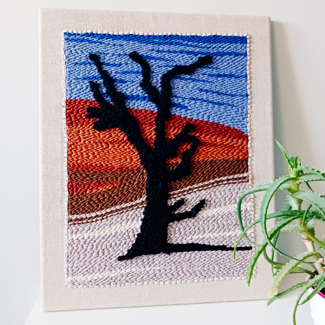 Dekoracyjny obraz wykonany techniką punch needle, przedstawiający czarne drzewo na tle pustynnego krajobrazu z czerwonymi wydmami i niebieskim niebem. Obraz oparty o ścianę, ustawiony na białej powierzchni obok doniczki z zieloną rośliną.