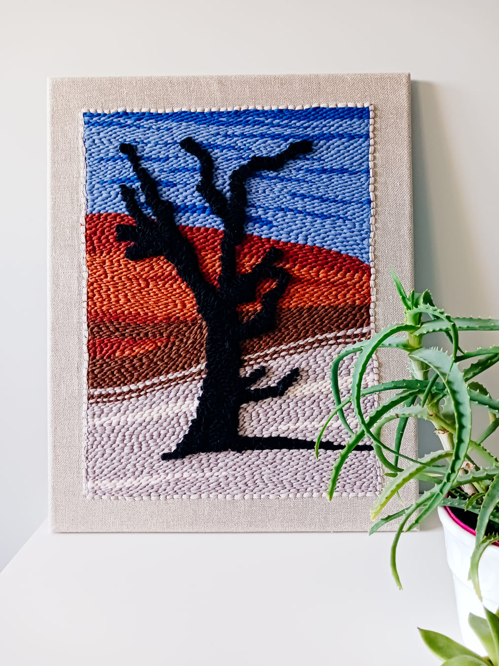 Dekoracyjny obraz wykonany techniką punch needle, przedstawiający czarne drzewo na tle pustynnego krajobrazu z czerwonymi wydmami i niebieskim niebem. Obraz oparty o ścianę, ustawiony na białej powierzchni obok doniczki z zieloną rośliną.