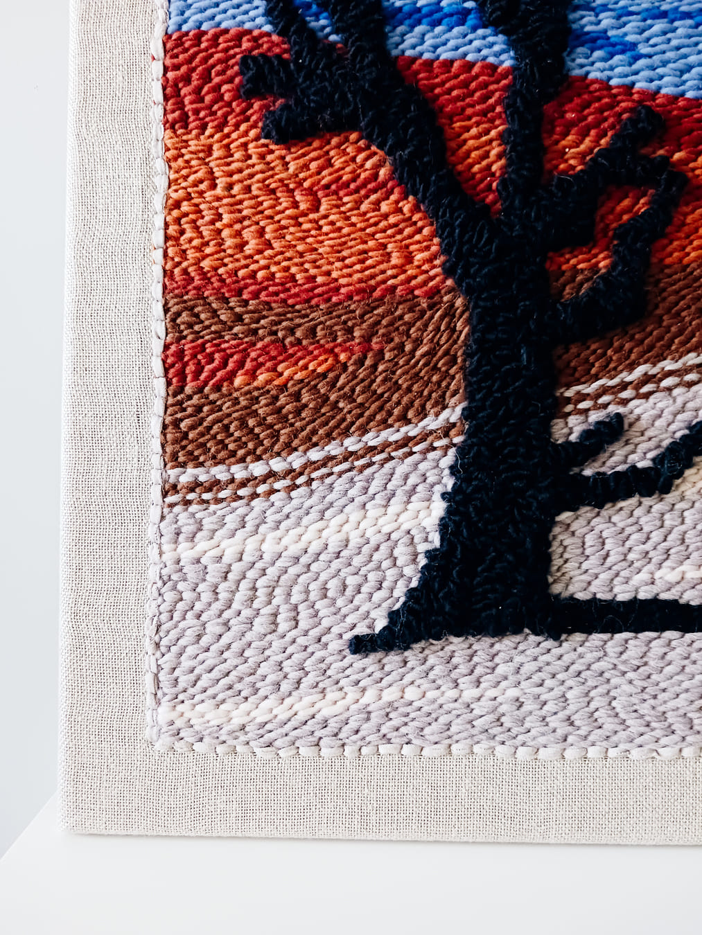 Fragment obrazu wykonanego techniką punch needle, który przedstawia czarne drzewo na tle pustynnego krajobrazu z czerwonymi wydmami i niebieskim niebem. Obraz oparty o ścianę, ustawiony na białej powierzchni.