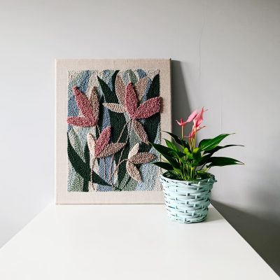 Teksturowany obraz wykonany techniką punch needle, przedstawiający abstrakcyjne liście w odcieniach różu i beżu na tle w kolorach zieleni i niebieskiego, stoi na białej powierzchni. Obok znajduje się doniczka z jasnozielonymi liśćmi i różowymi kwiatami, umieszczona w jasnoniebieskim, plecionym koszyku. Tło stanowi jasnoszara ściana.