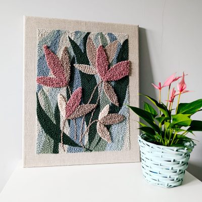 Teksturowany obraz wykonany techniką punch needle, przedstawiający abstrakcyjne liście w odcieniach różu i beżu na tle w kolorach zieleni i niebieskiego, stoi na białej powierzchni. Obok znajduje się doniczka z jasnozielonymi liśćmi i różowymi kwiatami, umieszczona w jasnoniebieskim, plecionym koszyku. Tło stanowi jasnoszara ściana.