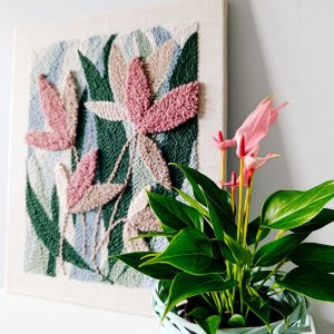 Teksturowany obraz wykonany techniką punch needle, przedstawiający abstrakcyjne liście w odcieniach różu i beżu na tle w kolorach zieleni i niebieskiego, stoi na białej powierzchni. Na pierwszym planie znajduje się doniczka z jasnozielonymi liśćmi i różowymi kwiatami, umieszczona w jasnoniebieskim, plecionym koszyku. Tło stanowi jasnoszara ściana.