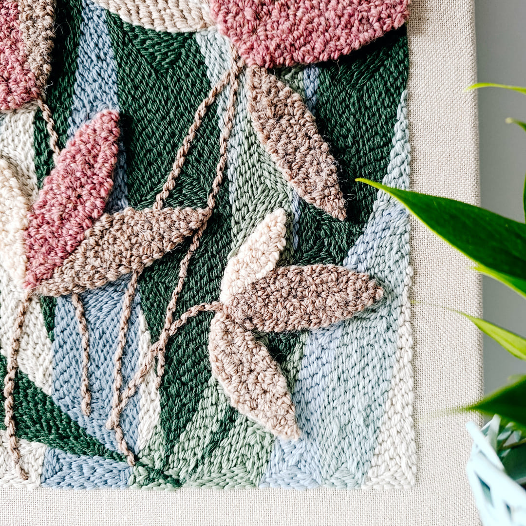 Fragment teksturowanego obrazu wykonanego techniką punch needle. Obraz przedstawia abstrakcyjne liście w odcieniach różu i beżu na tle w kolorach zieleni i niebieskiego.