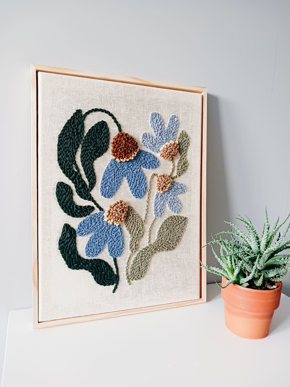 Ręcznie wykonany obraz techniką punch needle w odcieniach niebieskiego i zieleni oprawiony w wąską drewniana ramkę. Dekoracja ścienna o kwiatowym wzorze, idealna do nowoczesnych i przytulnych wnętrz.