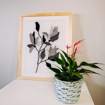 Haftowany obraz przedstawiający kwiatowy motyw w odcieniach szarości na białym tle, oprawiony w jasną drewnianą ramę. Dekoracja ścienna pasująca do eleganckich i minimalistycznych wnętrz.