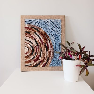 Obraz wykonany techniką punch needle, przedstawiający abstrakcyjny wzór wiru w odcieniach brązu, beżu i błękitu, oparty o ścianę na białej powierzchni. Obok stoi biała doniczka z rośliną o zielono-fioletowych liściach.