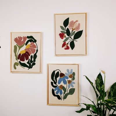 Trzy haftowane obrazki z motywem kwiatów powieszone niesymetrycznie na białej ścianie