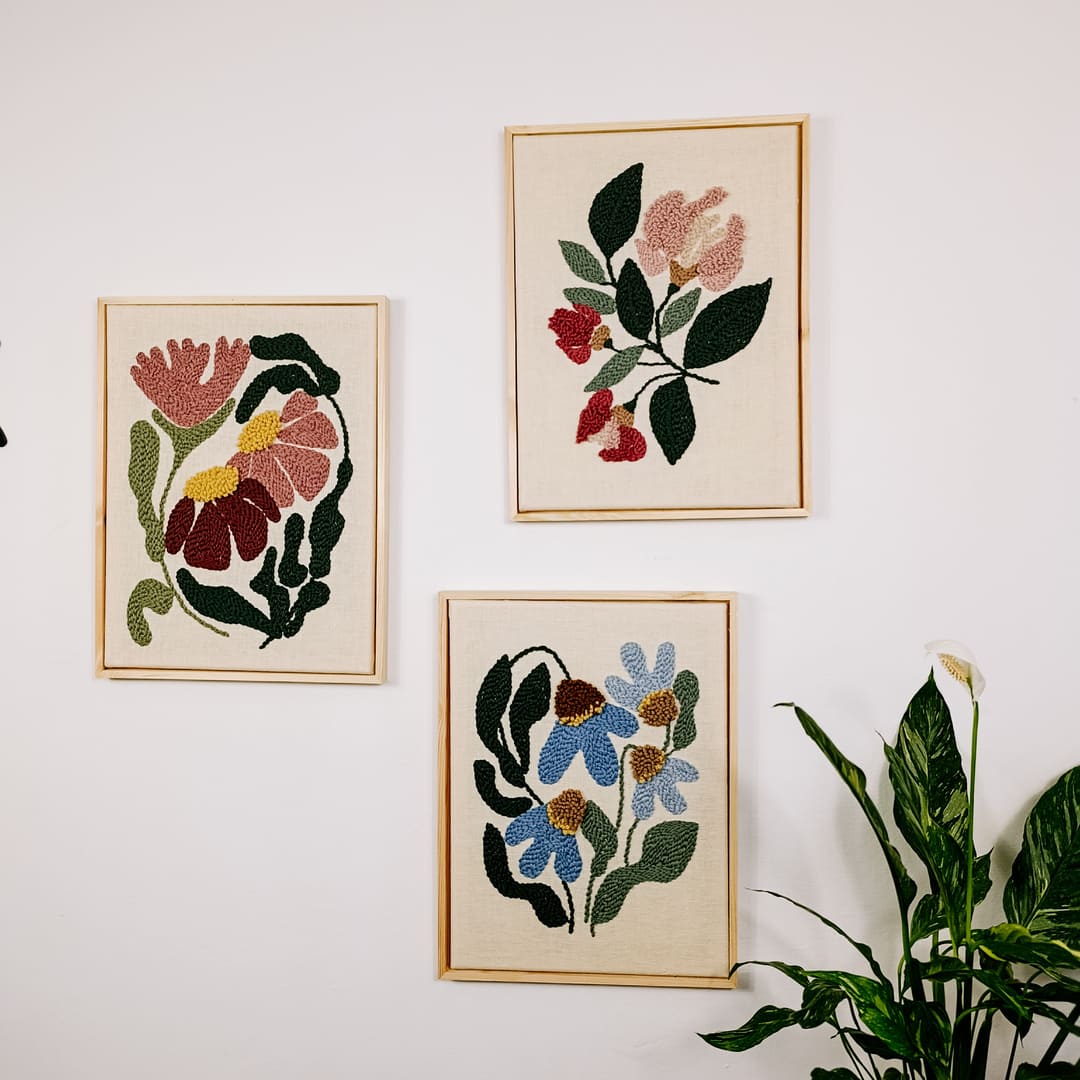 Trzy haftowane obrazki z motywem kwiatów powieszone niesymetrycznie na białej ścianie