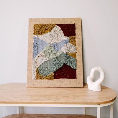Obraz z wełny w abstrakcyjne, geometryczne formy. Zróżnicowane faktury i kontrastujące kolory. Stoi na stoliku, oparty o jasną ścianę, obok biały wazonik w abstrakcyjnym kształcie.