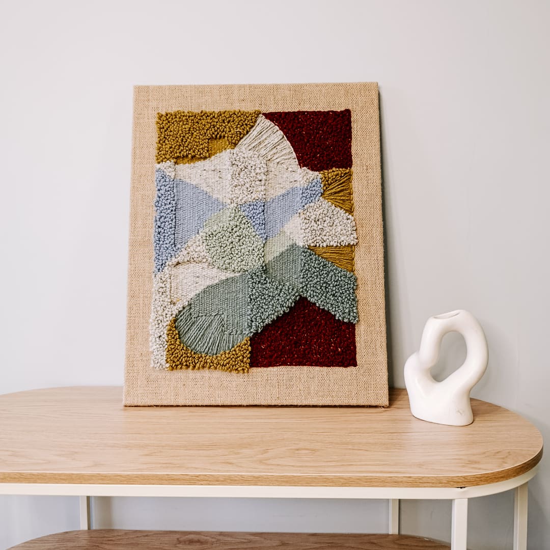 Obraz z wełny w abstrakcyjne, geometryczne formy. Zróżnicowane faktury i kontrastujące kolory. Stoi na stoliku, oparty o jasną ścianę, obok biały wazonik w abstrakcyjnym kształcie.