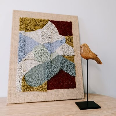 Obraz z wełny w abstrakcyjne, geometryczne formy. Zróżnicowane faktury i kontrastujące kolory. Stoi na stoliku, oparty o jasną ścianę, obok drewniana figurka ptaszka na czarnej nóżce.