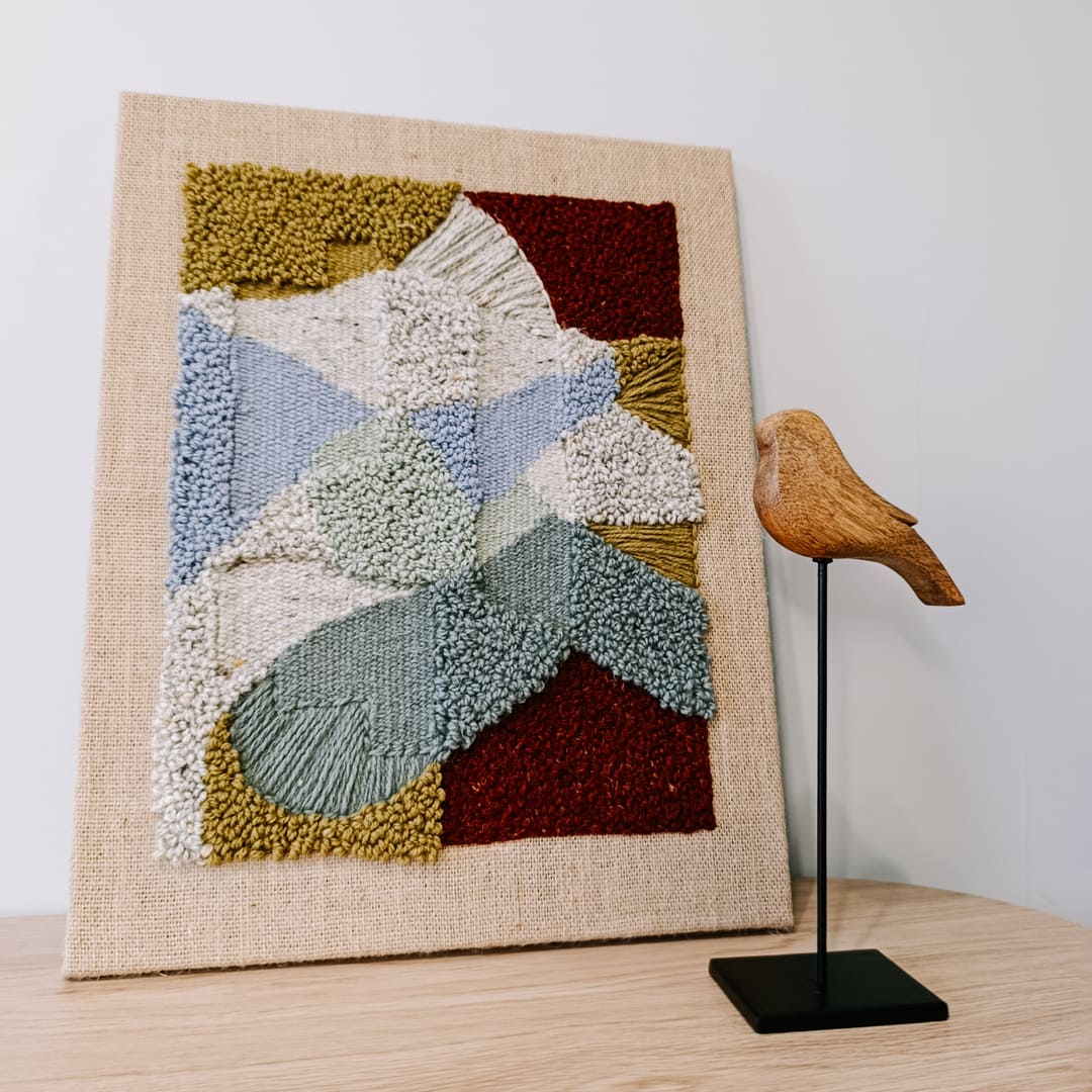 Obraz z wełny w abstrakcyjne, geometryczne formy. Zróżnicowane faktury i kontrastujące kolory. Stoi na stoliku, oparty o jasną ścianę, obok drewniana figurka ptaszka na czarnej nóżce.
