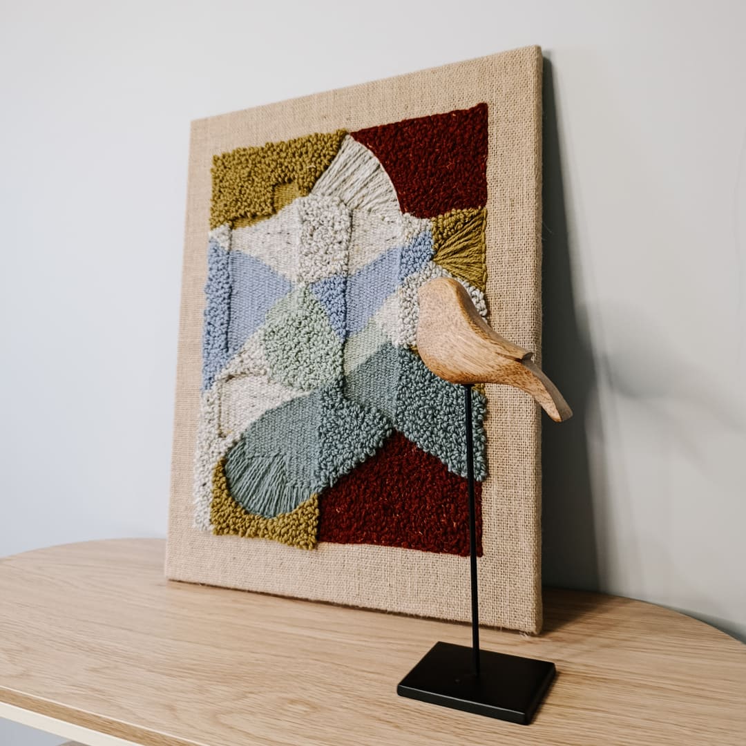 Obraz z wełny w abstrakcyjne, geometryczne formy. Zróżnicowane faktury i kontrastujące kolory. Stoi na stoliku, oparty o jasną ścianę, obok drewniana figurka ptaszka na czarnej nóżce.