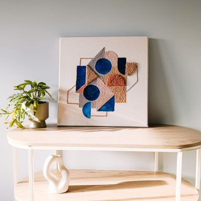 Obraz abstrakcyjny o geometrycznej kompozycji w technice mix media - malowanie stemplami i haft punch needle. Stoi na półce oparty o jasną ścianę, obok kwiat w doniczce i wazonik o nieregularnym kształcie.