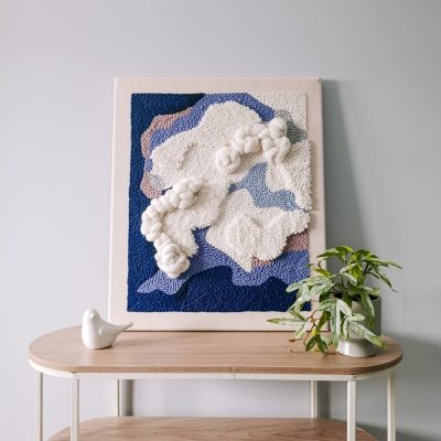 Trójwymiarowy obraz punch needle w niebiesko-białej kolorystyce. Abstrakcyjna kompozycja. Obraz stoi na półce, oparty o jasnoszarą ścianę. Obok kwiat w doniczce i ceramiczna figurka ptaszka.