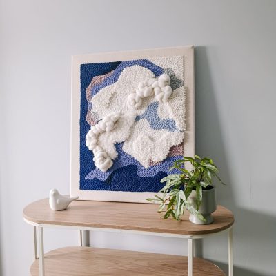 Trójwymiarowy obraz punch needle w niebiesko-białej kolorystyce. Abstrakcyjna kompozycja. Obraz stoi na półce, oparty o jasnoszarą ścianę. Obok kwiat w doniczce i ceramiczna figurka ptaszka.