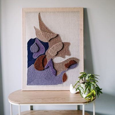 Abstrakcyjny obraz tekstylny w kolorach fioletu i jasnych brązów. Stoi na półce oparty o ścianę. Obok kwiat w doniczce i ceramiczna figurka ptaszka.