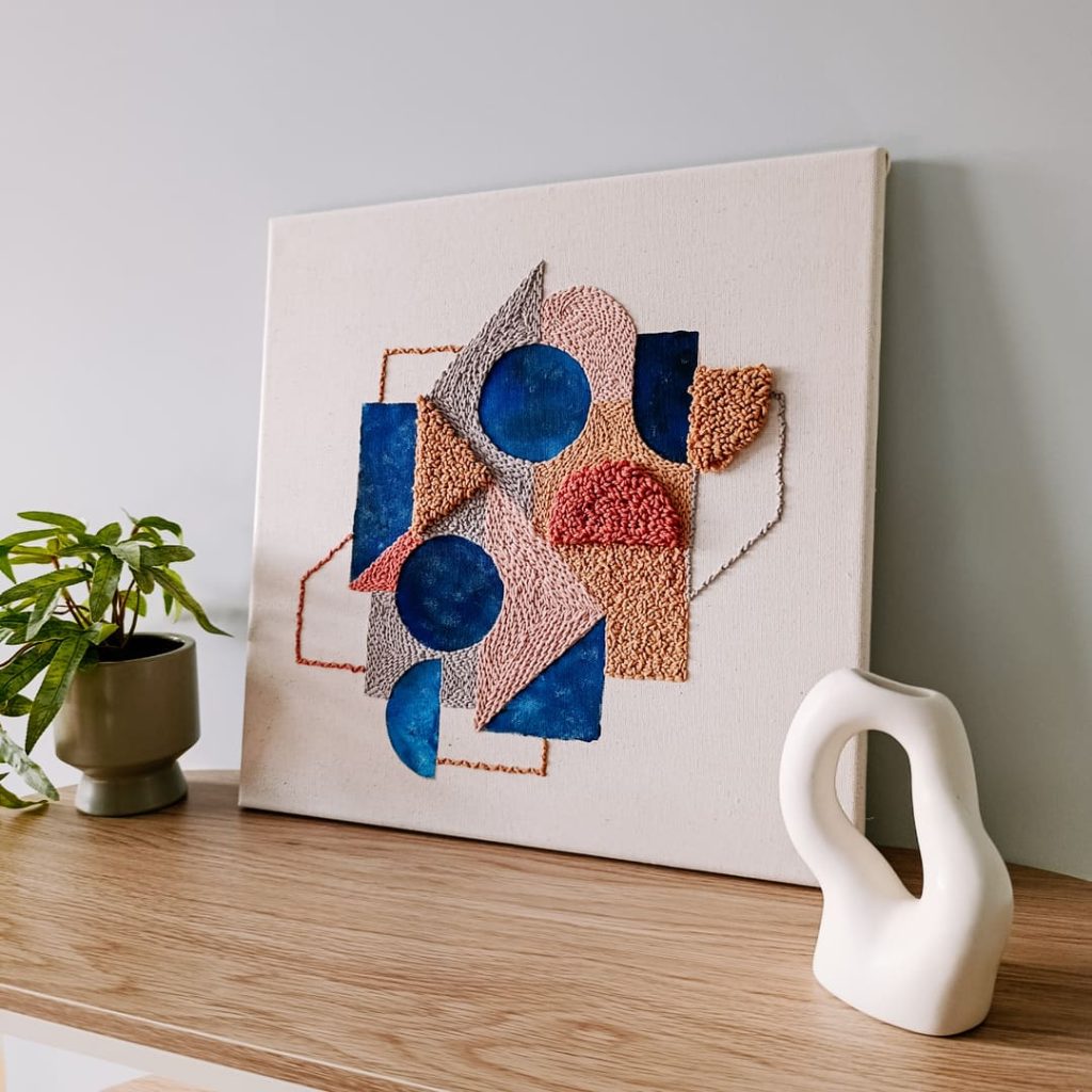 Geometryczny obraz punch needle w pastelowych kolorach z elementami stemplowania płótna farbą. 