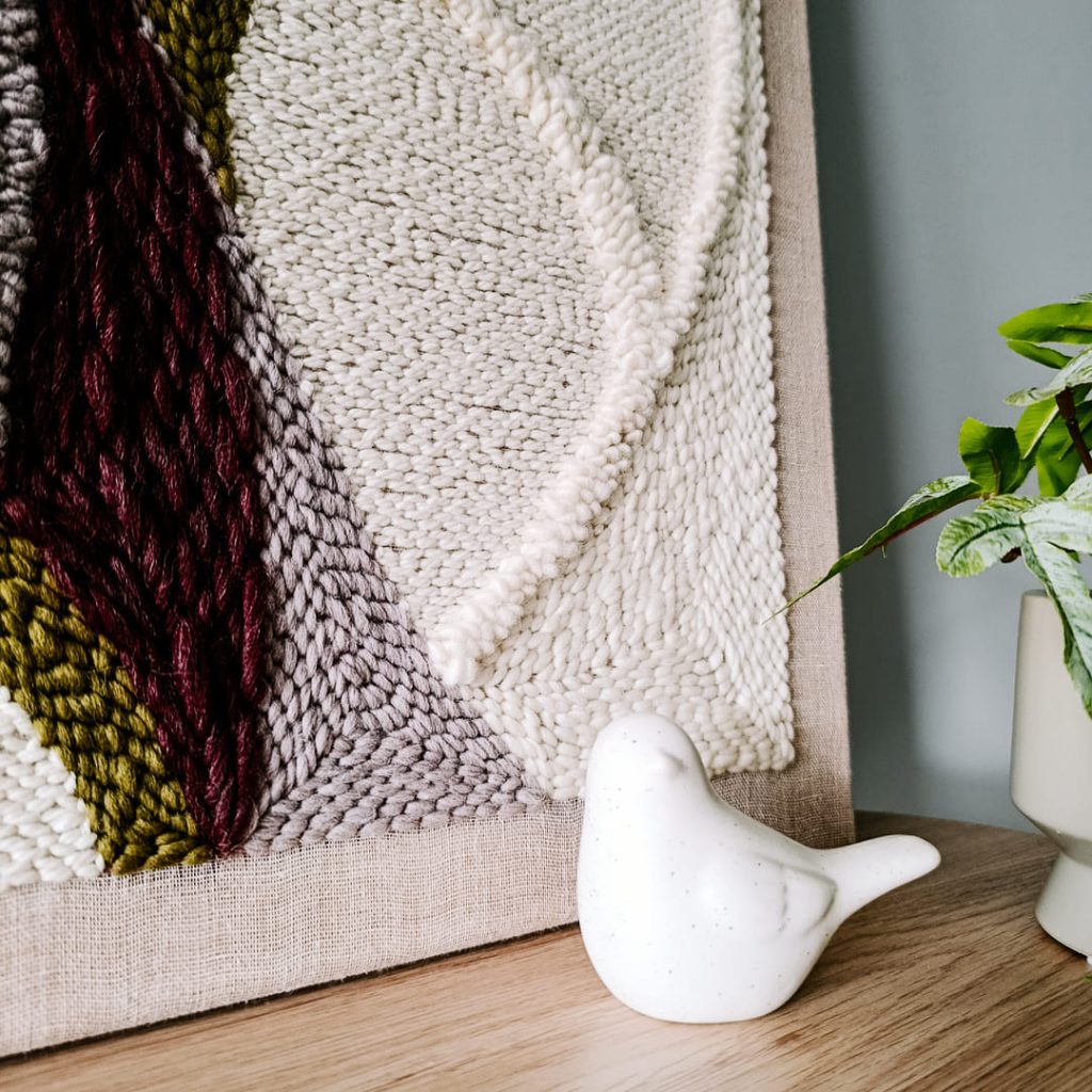 Fragment obrazu punch needle w kolorach ziemi. Obok mały ceramiczny ptaszek.